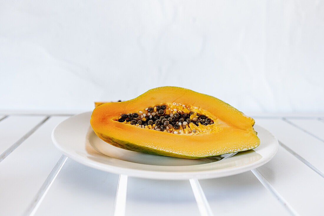 Halbierte frische reife exotische Papaya mit Kernen, serviert auf einem Keramikteller auf einem weißen Tisch an einem sonnigen Sommertag
