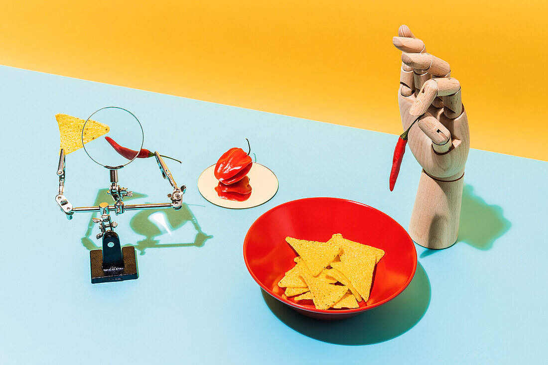 From above magnifying glass and artificial hand placed near hot peppers and bowl with tortilla chips for Mexican cuisine dish on yellow and blue background