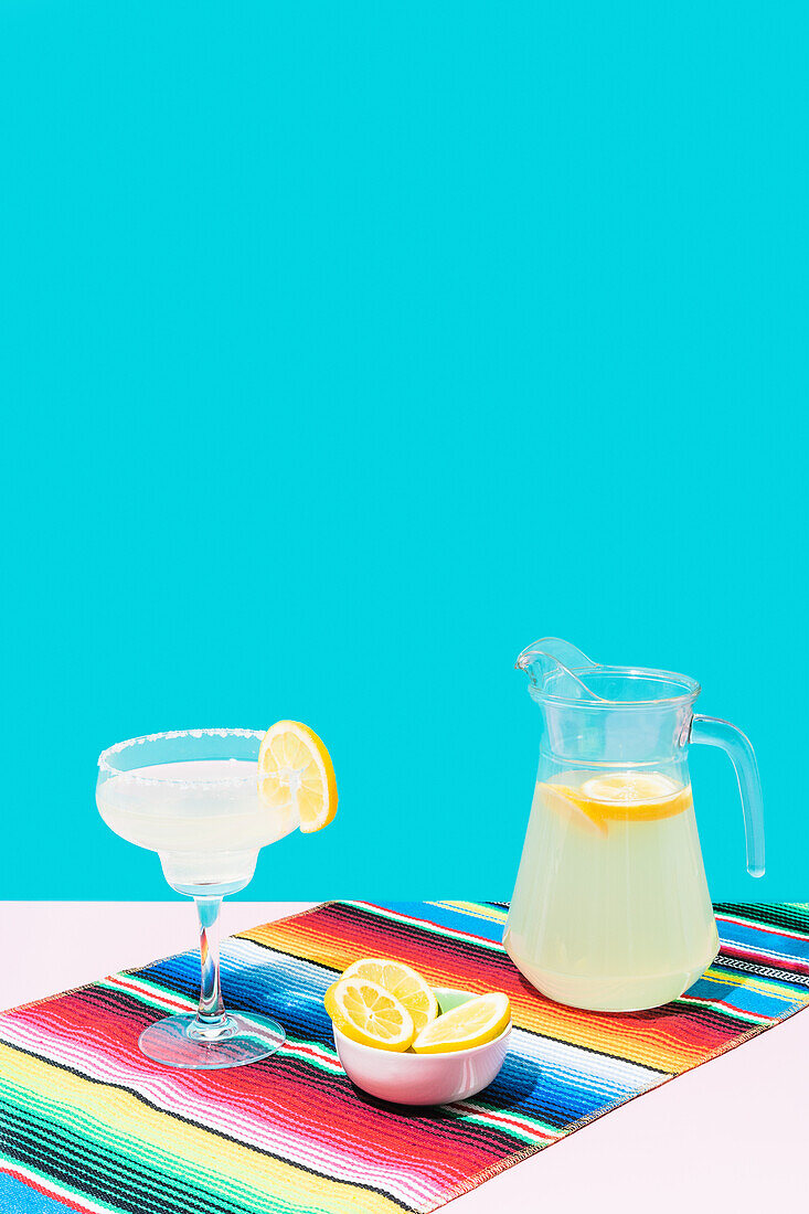Glass and jug of cold lemonade with fresh lemons placed on colorful Mexican rug blanket on blue background
