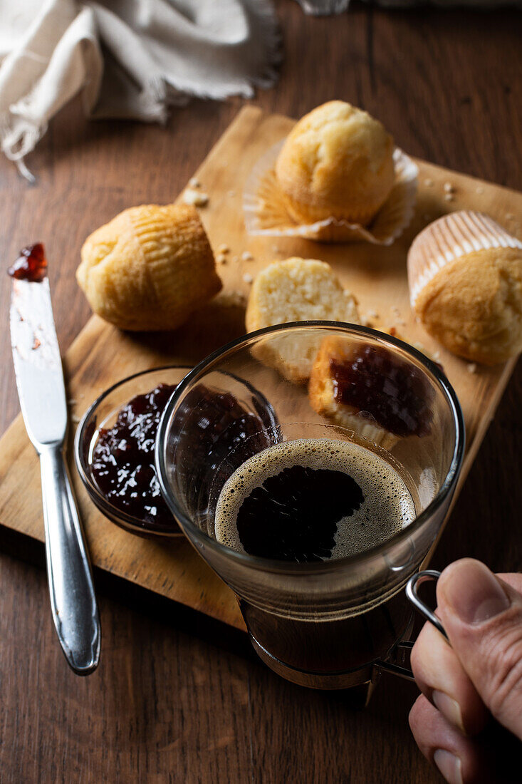 Abgeschnittene unerkennbare Person, die einen Becher mit Kaffee hält und leckere hausgemachte Muffins mit Himbeermarmelade auf einem Holzbrett auf dem Tisch isst