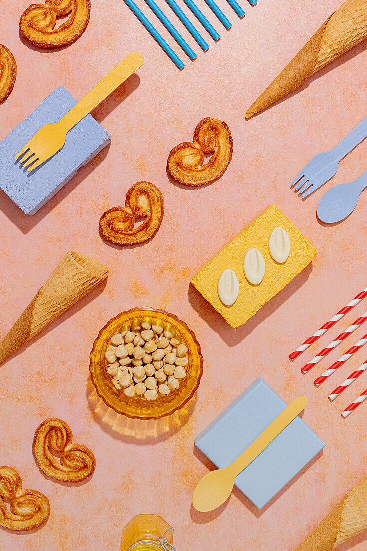 Overhead view of waffle cones and elephant ear cookies with candy placed against disposable cutlery and straw on orange pastel backdrop