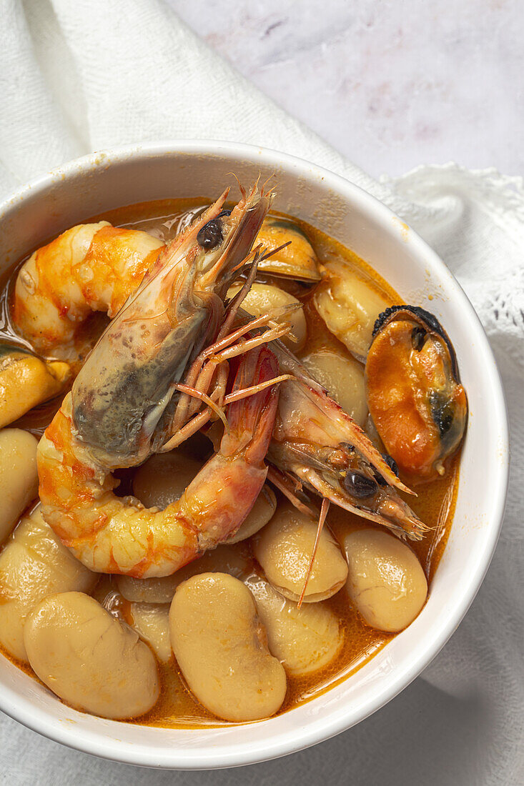 From above bowl with typical stew of beans with prawns, shrimps and mussels on a lace tablecloth and on a stone table