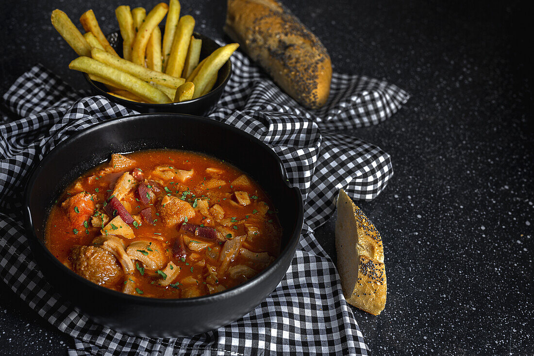 From above of tasty callos a la madrilena in black bowl near French fries and bread with seeds