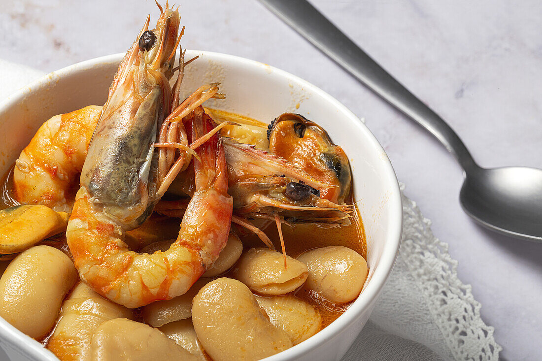 Crop of bowl with typical stew of beans with prawns, shrimps and mussels on a lace tablecloth close to a spoon and on a stone table