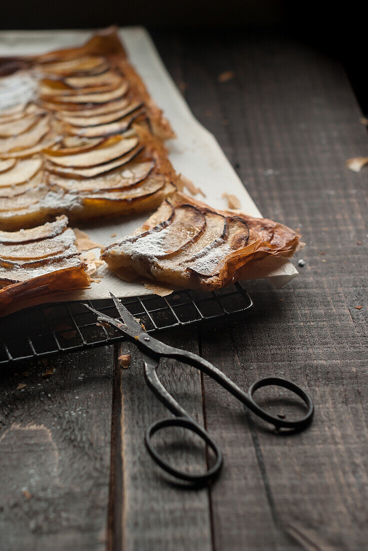 Closeup of cooling rack with frangipane apple pie on parchment lying on wooden table with old scissors