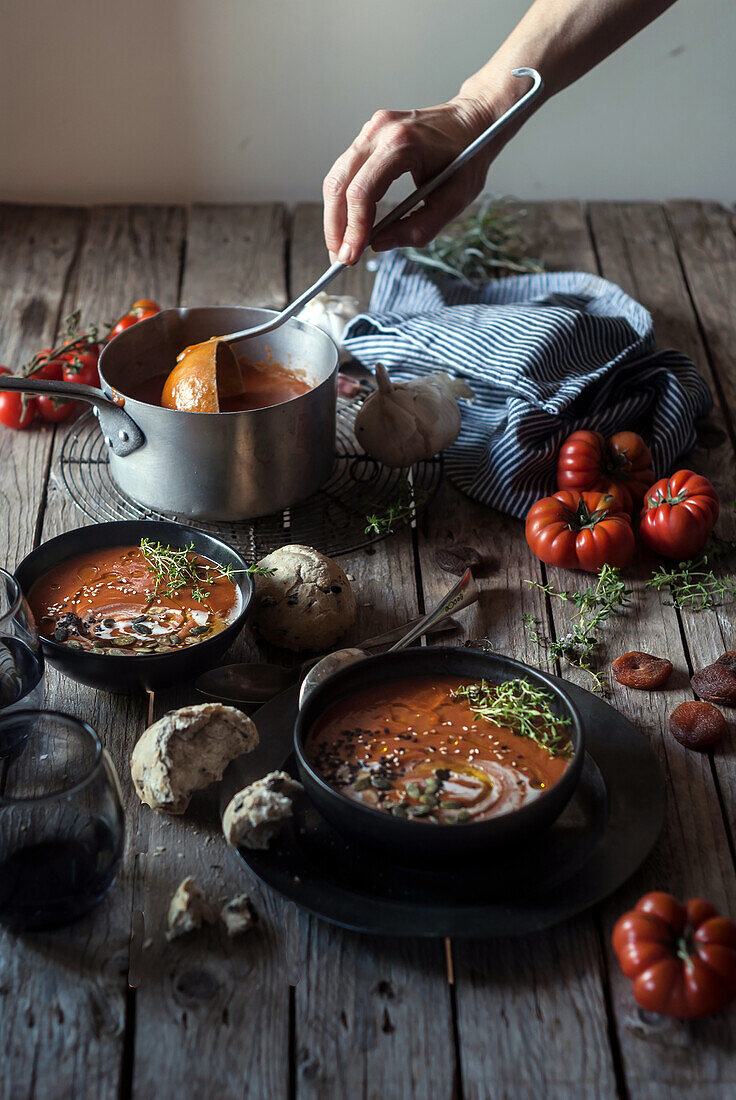 Eine Person hält eine Suppenkelle und serviert eine cremige Tomatensuppe mit Samen auf einem Tisch mit getrockneten Aprikosen und Brot
