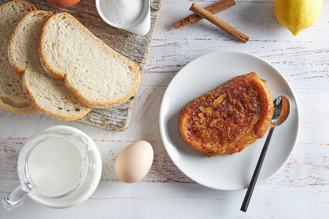 From above plate of appetizing Spanish torrija bread placed on table with raw eggs and sugar near wooden cutting board with jug of fresh milk and bread slices