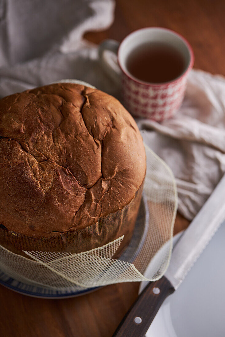 High angle of uncut fresh baked artisan Christmas panettone cake on wooden table next to cup of tea