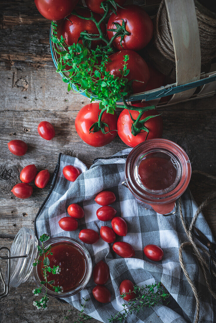 Draufsicht auf reife Tomaten und grüne Kräuter in einem Korb neben einer karierten Serviette und Gläsern mit Marmelade auf einem Küchentisch aus Holz