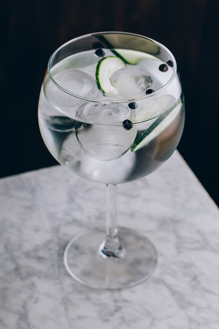 Glass of refreshing alcohol gin tonic cocktail with sliced cucumber and ice cubes on table