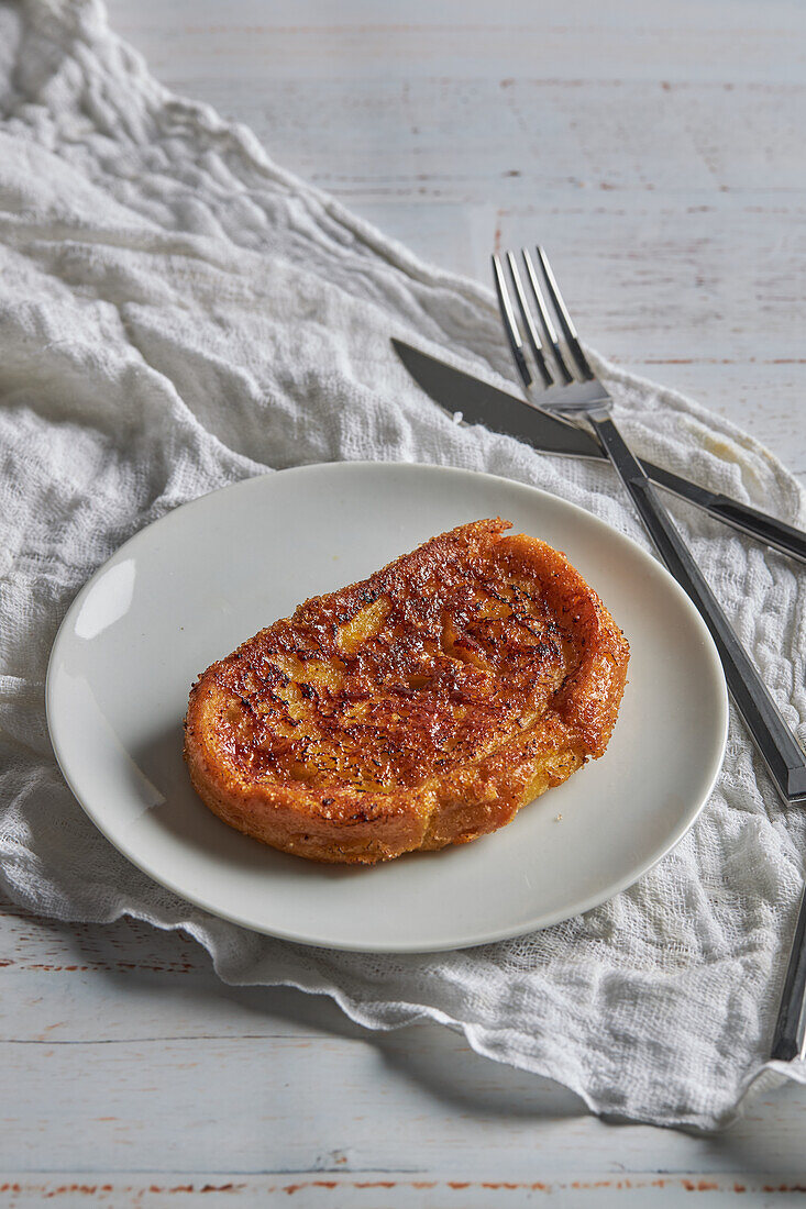 Blick von oben auf eine appetitliche, knusprige Torrija, die auf einem Teller angerichtet und auf einem weißen Holztisch mit Gabel und Messer serviert wird