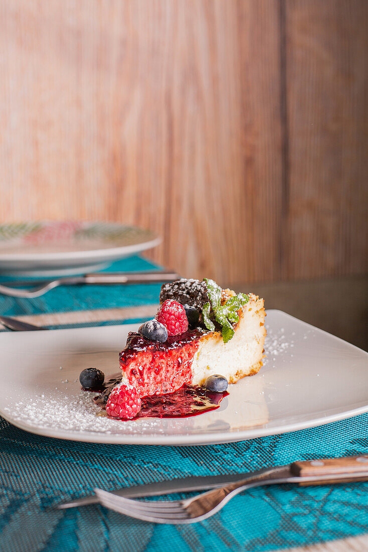 Tasty cheesecake with red jam, ripe raspberries, blueberries, fresh mint leaves and powdered sugar in ceramic white plate in restaurant