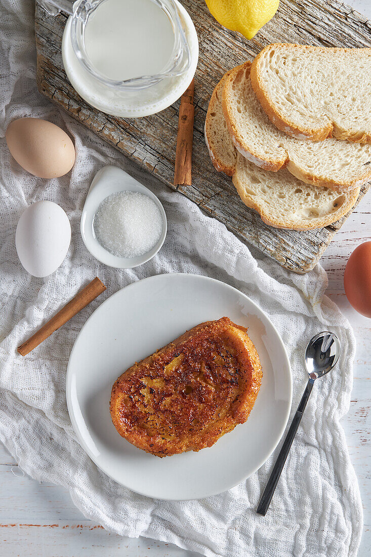 Blick von oben auf einen Teller mit appetitlichem spanischem Torrija-Brot, das mit rohen Eiern und Zucker auf dem Tisch liegt, neben einem hölzernen Schneidebrett mit einem Krug frischer Milch und Brotscheiben