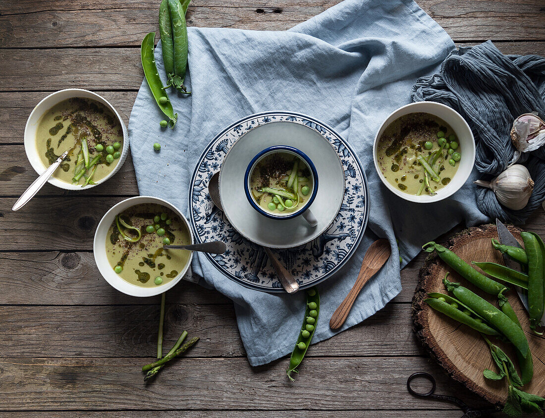 Flat lay of bowls with green pea and coconut cream soup on wooden table with pea pods and garlic in composition