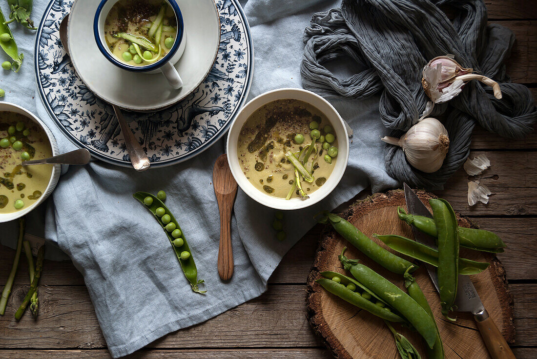 Flat lay of bowls with green pea and coconut cream soup on wooden table with pea pods and garlic in composition