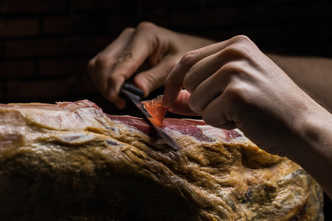 Crop unrecognizable cook cutting slice of tasty dry cured Spanish jamon on black background
