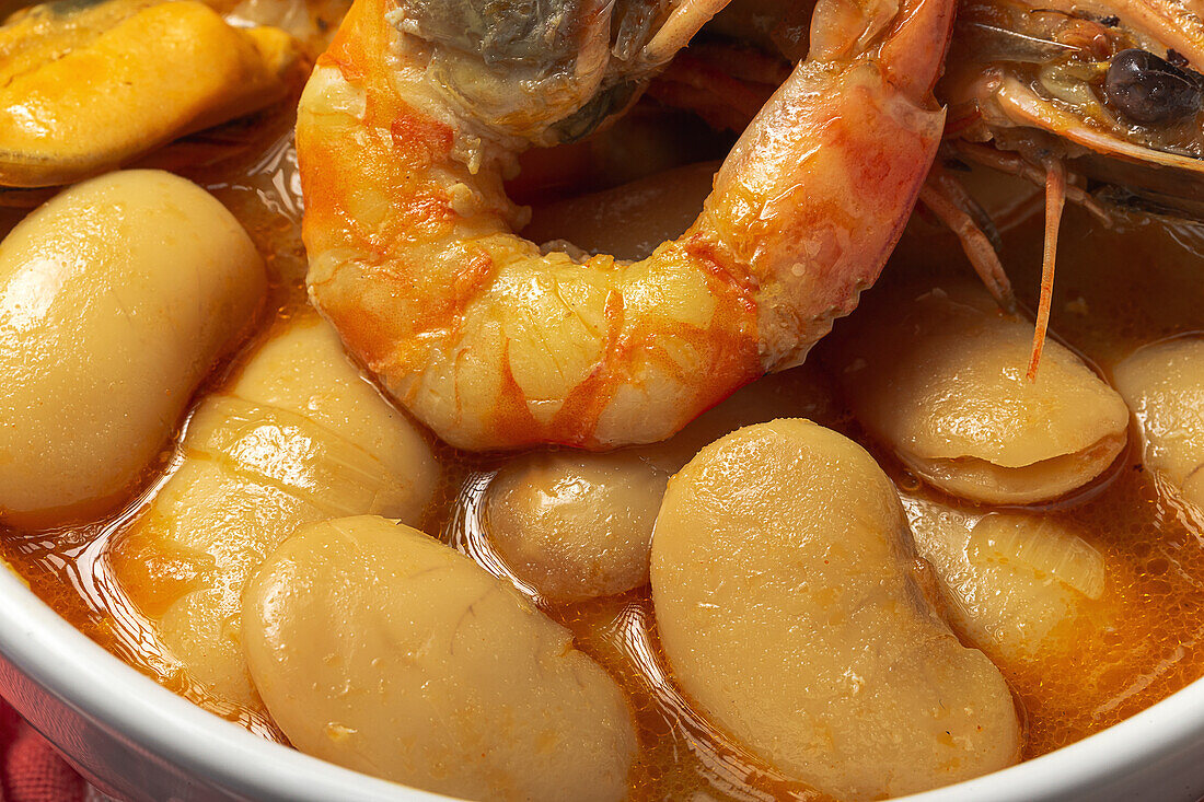 Close up of bowl with typical stew of beans with prawns, shrimps and mussels on a red checkered on a stone table