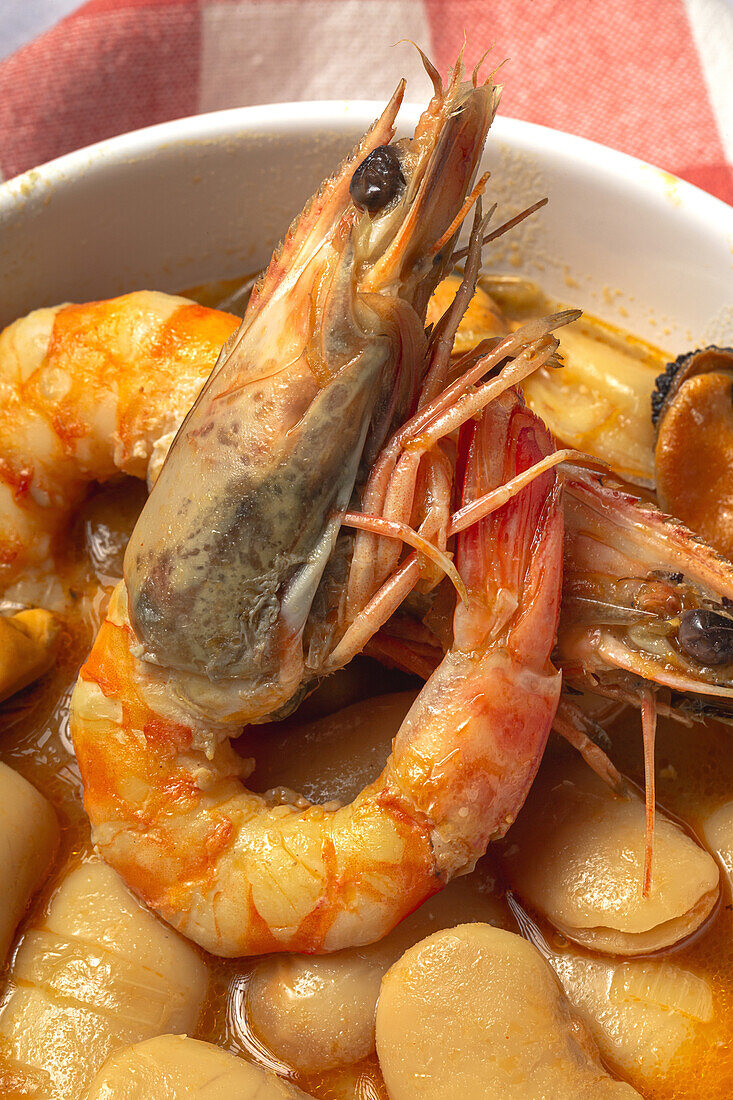 Nahaufnahme einer Schüssel mit einem typischen Bohneneintopf mit Garnelen, Krabben und Muscheln auf einem rot karierten Steintisch