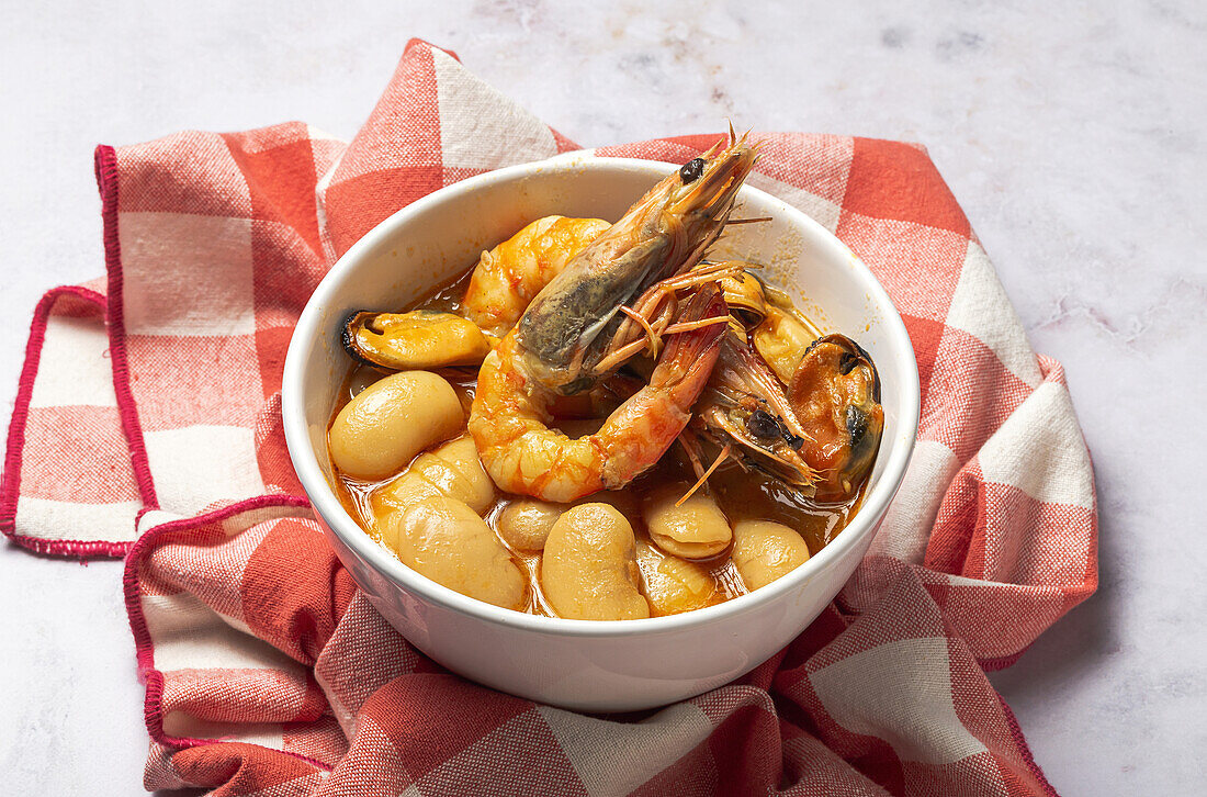 From above bowl with typical stew of beans with prawns, shrimps and mussels on a red checkered on a stone table