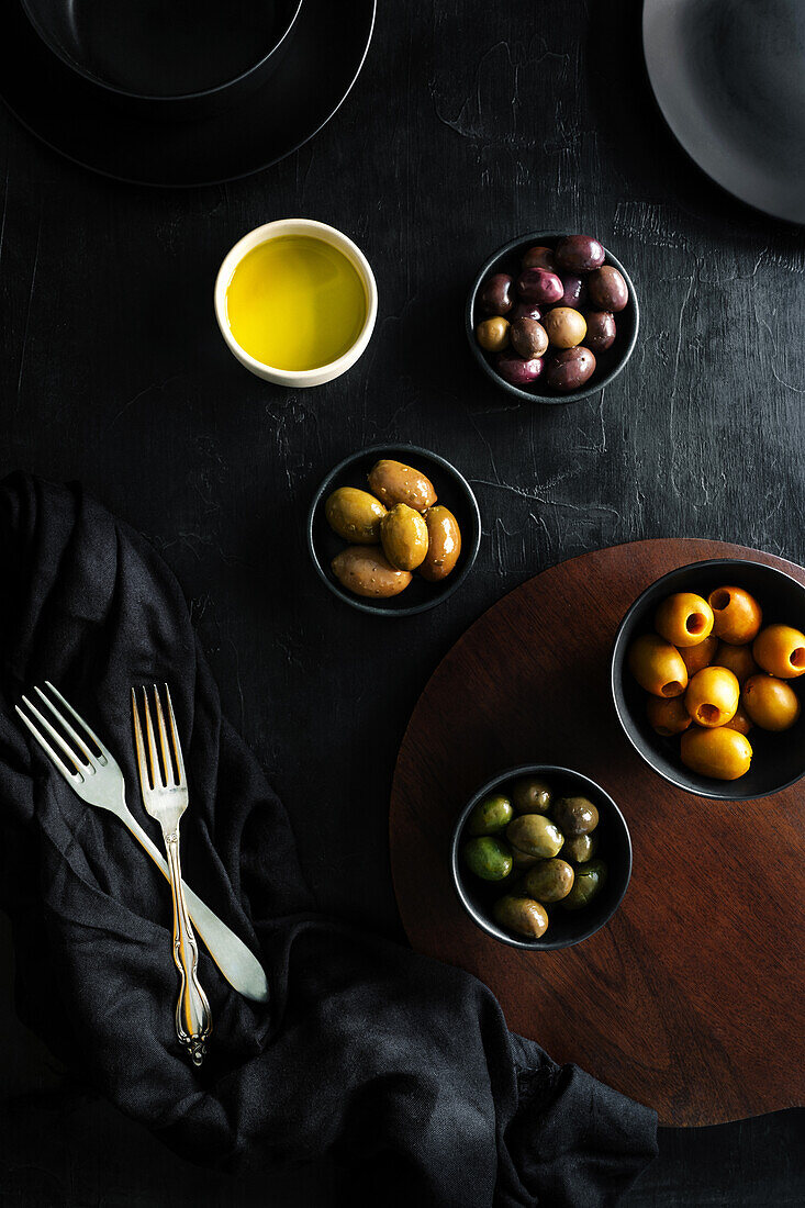 From above tasty juicy green brown yellow olives and oil in black and white bowls on black table decorated with tablecloth and forks