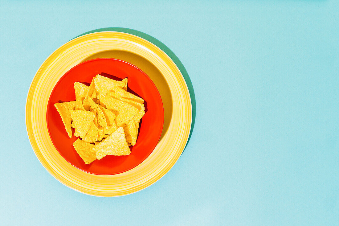 Top view of crispy tortilla chips placed on red plate near bowl with tasty guacamole sauce on blue and yellow background