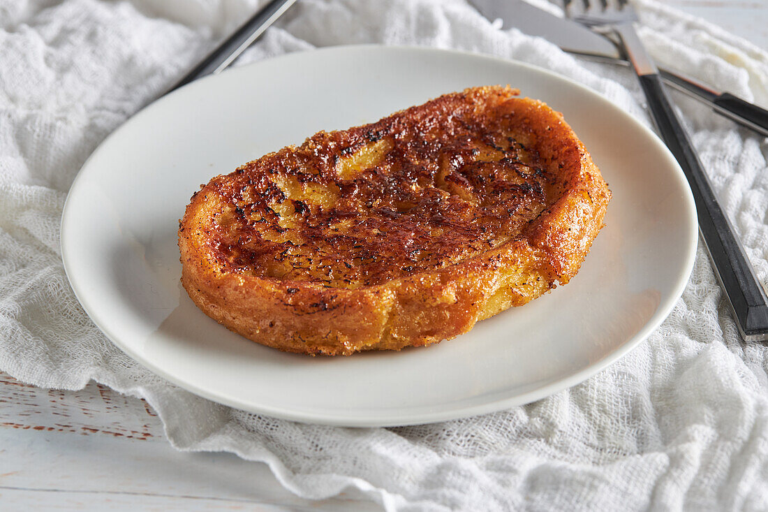 Blick von oben auf eine appetitliche, knusprige Torrija, die auf einem Teller angerichtet und auf einem weißen Holztisch mit Gabel und Messer serviert wird