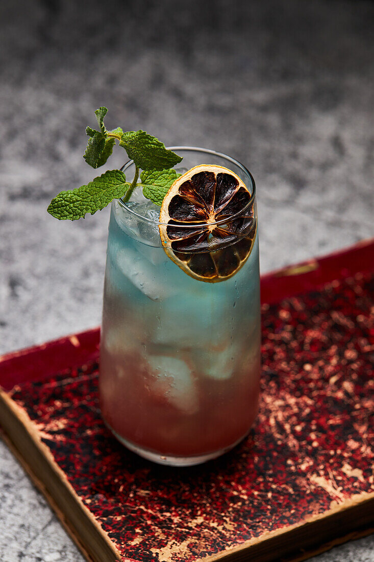 Glass of rainbow paradise colorful cocktail garnished with slice of orange and mint leaf on old book board on table