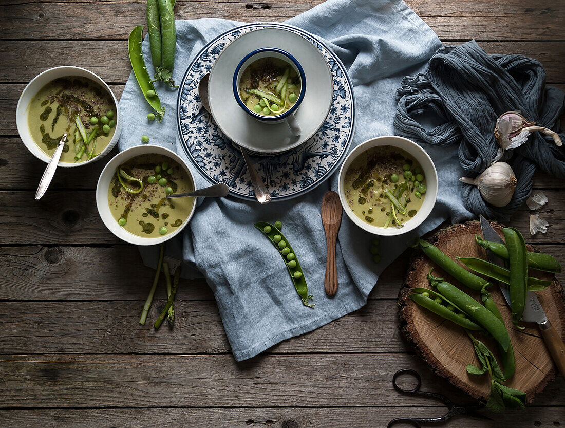 Flat lay of bowls with green pea and coconut cream soup on wooden table with pea pods and garlic in composition