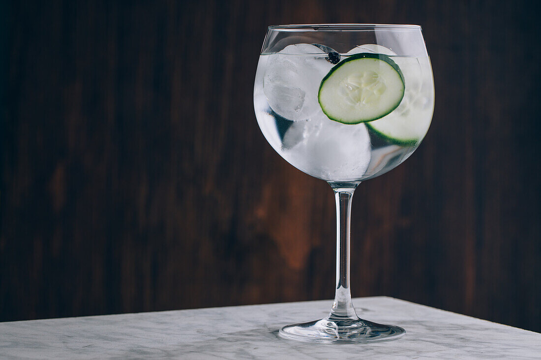 Glass of refreshing alcohol gin tonic cocktail with sliced cucumber and ice cubes on table