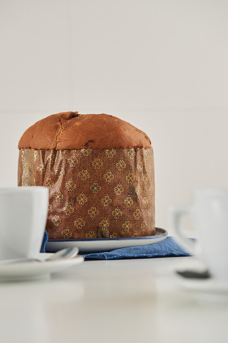 Ungeschnittener, frisch gebackener, handwerklich hergestellter Weihnachts-Panettone-Kuchen auf einem Teller, der auf einem blauen Tuch neben Tassen an einer weißen Wand steht