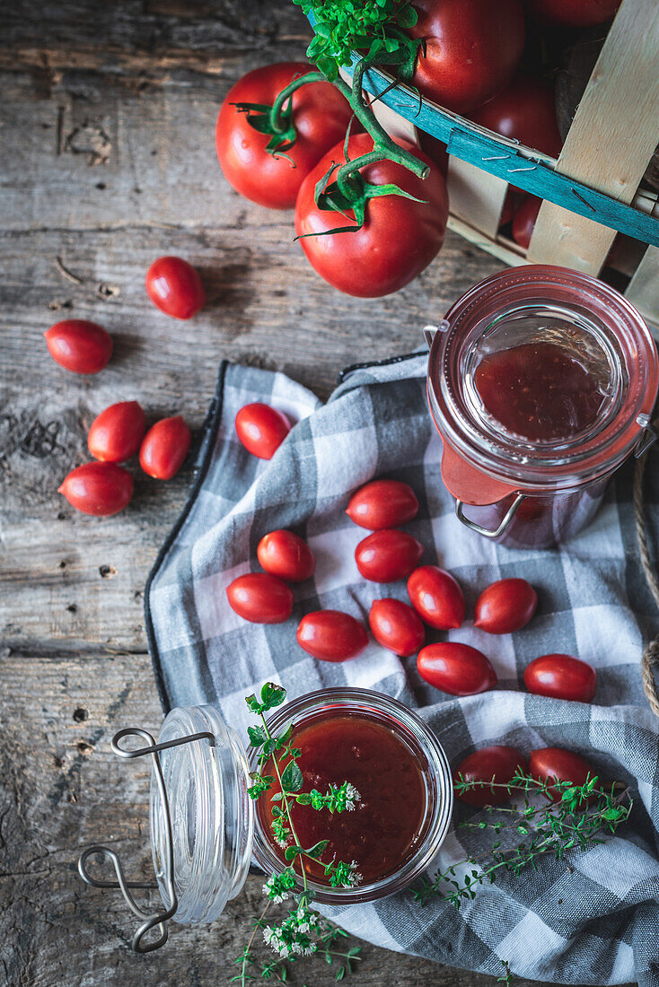 Draufsicht auf reife Tomaten und grüne Kräuter in einem Korb neben einer karierten Serviette und Gläsern mit Marmelade auf einem Küchentisch aus Holz