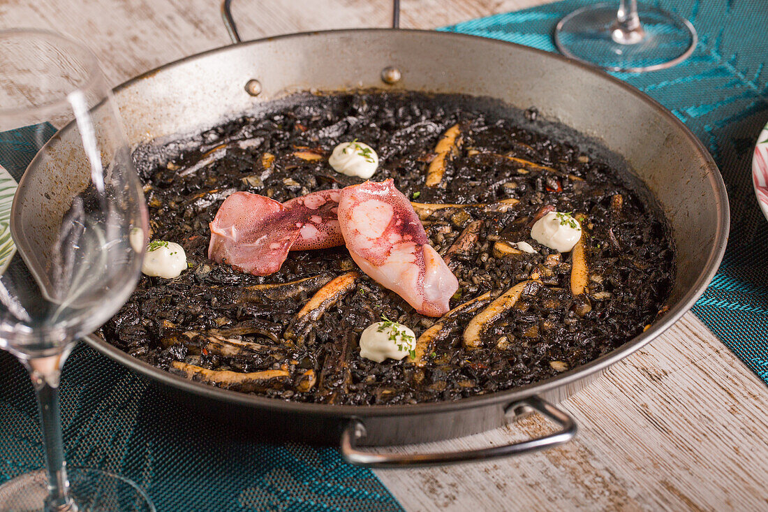 Hoher Blickwinkel auf köstlichen schwarzen Reis und Tintenfisch garniert mit Aioli-Sauce, serviert in einer Metallpfanne auf einem Tisch in einem Restaurant