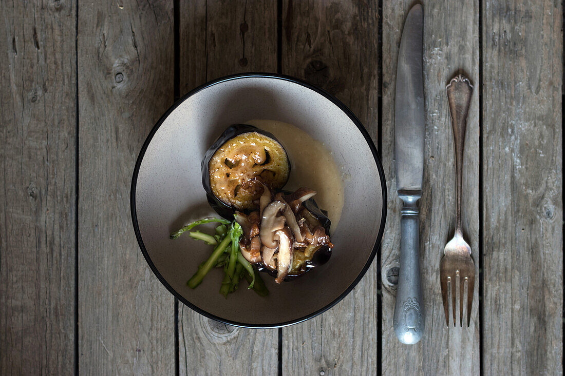 Servierte runde Schale mit Auberginen und Shiitake-Pilzen auf Holztisch mit Silberbesteck von oben gesehen