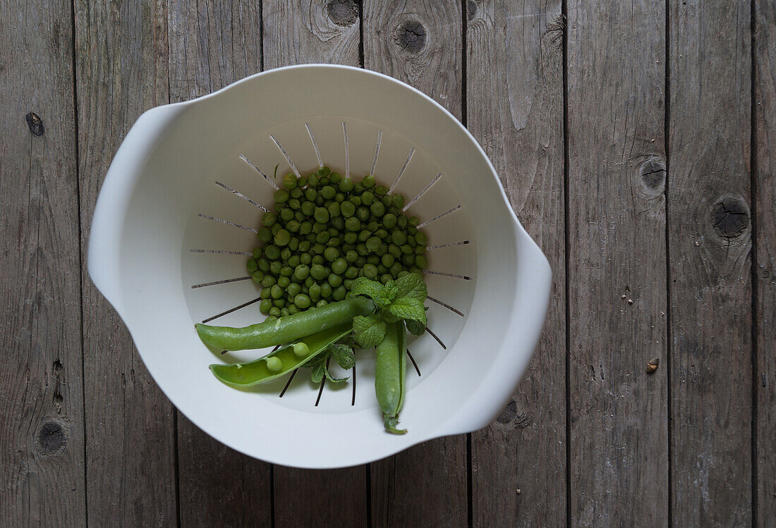 Top view of white plastic drain bowl full of peas and pods and fresh mint leaves on wooden table