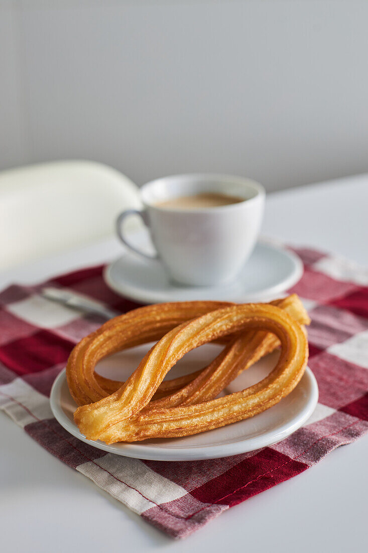 Appetitliche Churros auf einem Teller und eine Tasse frischen Morgenkaffee auf einer Serviette auf dem Frühstückstisch in der Küche