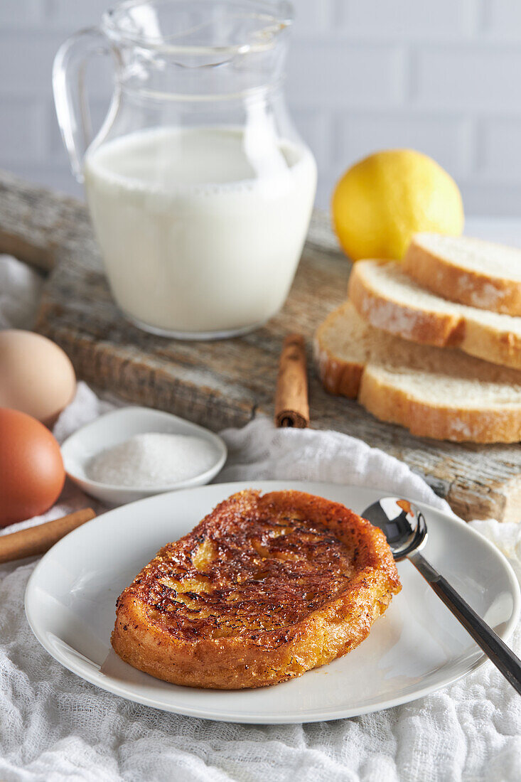 Blick von oben auf einen Teller mit appetitlichem spanischem Torrija-Brot, das mit rohen Eiern und Zucker auf dem Tisch liegt, neben einem hölzernen Schneidebrett mit einem Krug frischer Milch und Brotscheiben