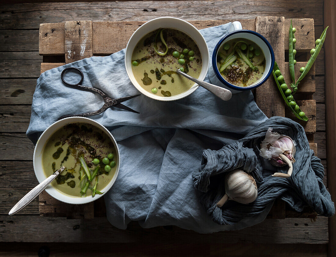 Flat lay of bowls with green pea and coconut cream soup on wooden table with pea pods and garlic in composition