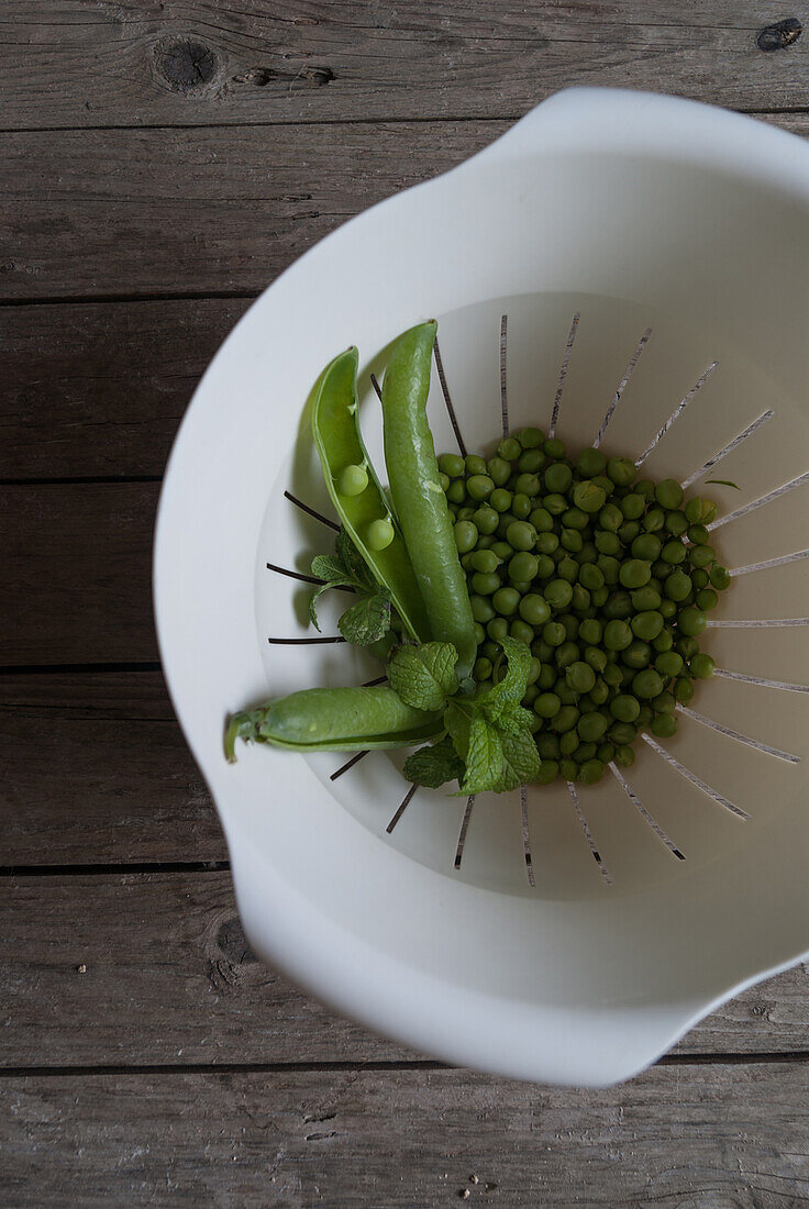 Top view of white plastic drain bowl full of peas and pods and fresh mint leaves on wooden table