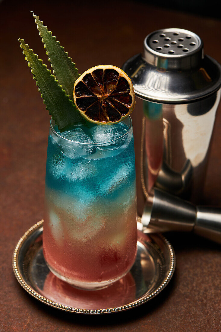 Glas Regenbogenparadies mit buntem Cocktail, garniert mit einer Orangenscheibe neben einem Jigger auf einem Metalltablett auf dem Tisch