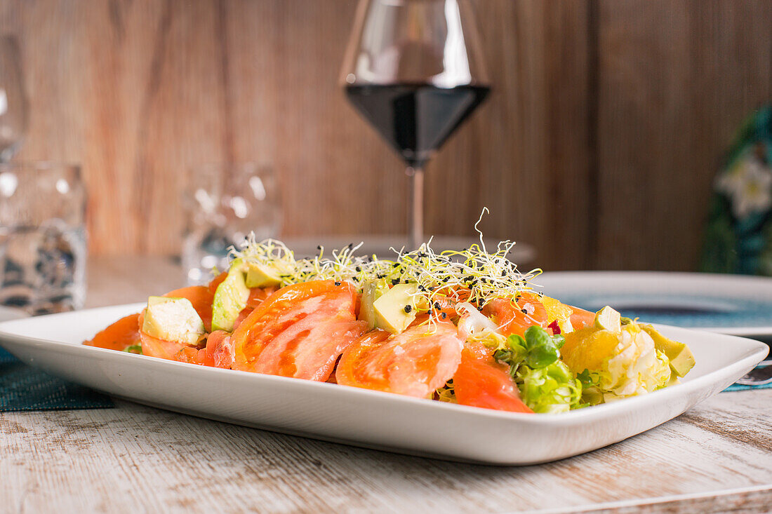 Köstlicher Salat mit geschnittenen frischen Tomaten und Avocado mit mariniertem Lachs unter Sprossen neben einem Glas Rotwein auf dem Tisch