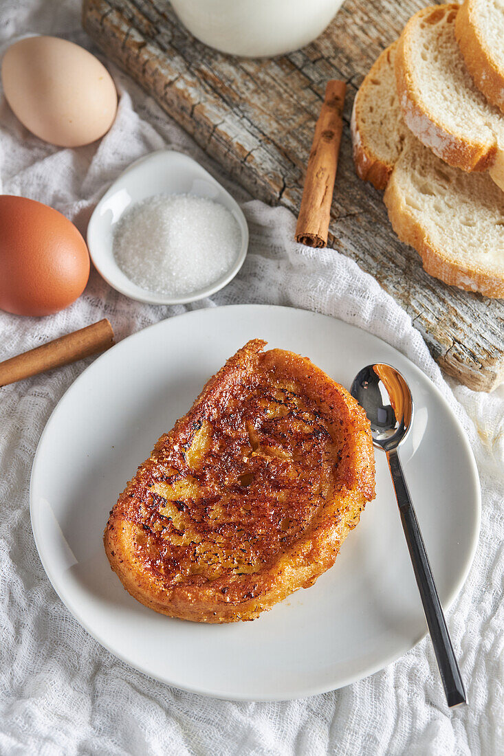 Blick von oben auf einen Teller mit appetitlichem spanischem Torrija-Brot, das mit rohen Eiern und Zucker auf dem Tisch liegt, neben einem hölzernen Schneidebrett mit einem Krug frischer Milch und Brotscheiben