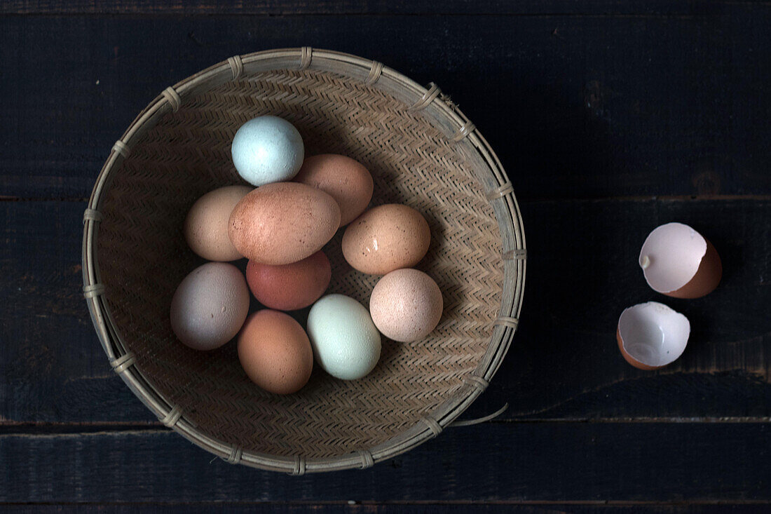 From above shot of wicker bowl filled with raw eggs on wooden table with eggshell near