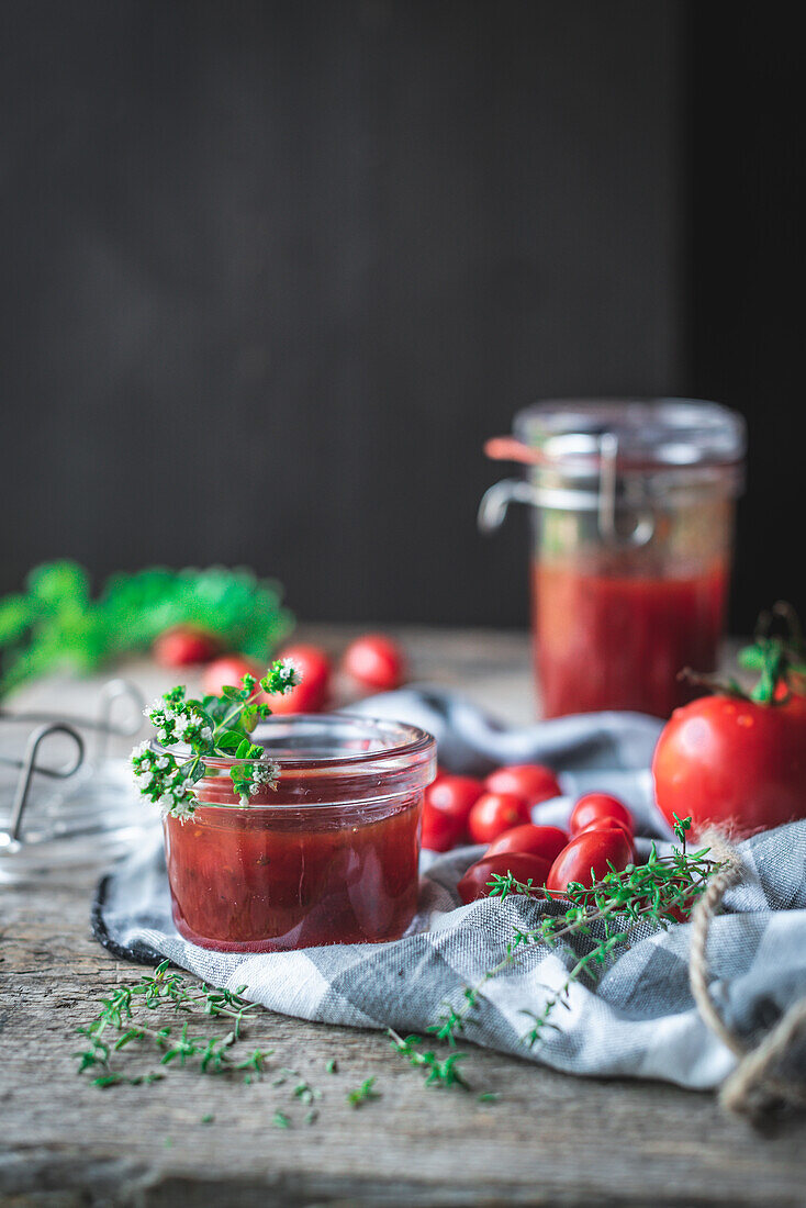 Soft focus of small glass jar with fresh tomato jam placed near checkered napkin and green herbs on wooden table