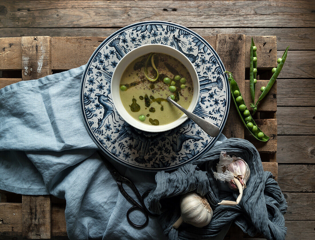 Flat lay of bowls with green pea and coconut cream soup on wooden table with pea pods and garlic in composition