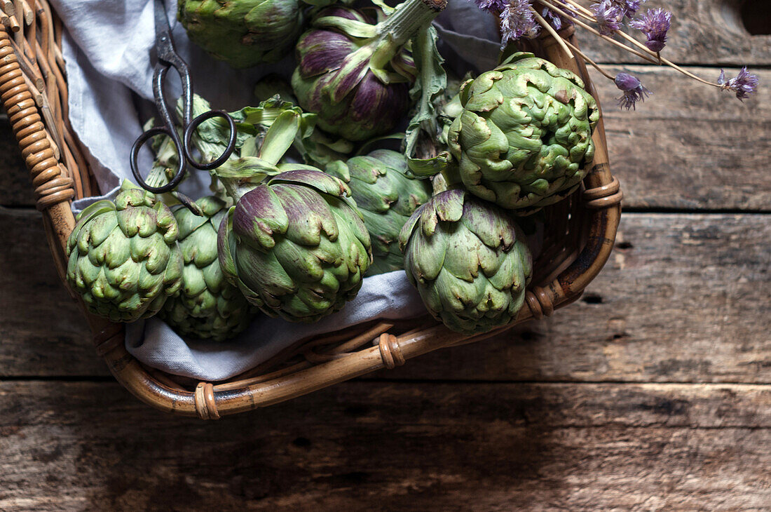 Closeup from above view of green artichokes laid in wicker basket with bunch of little purple flowers