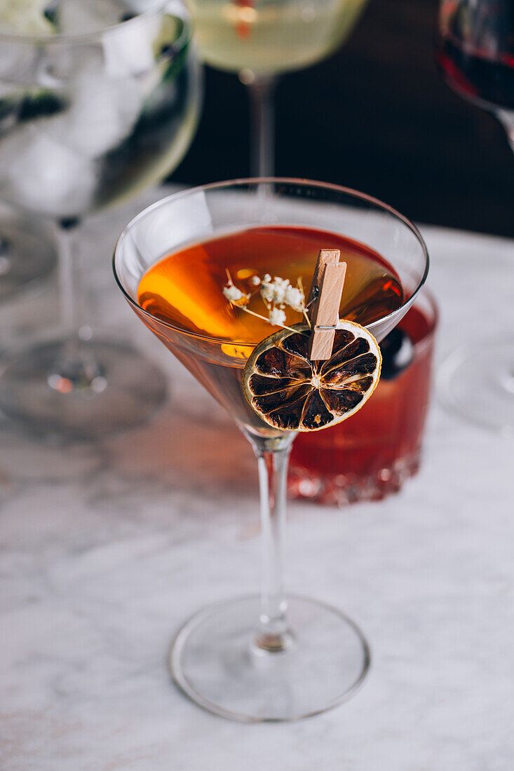 Glass of traditional alcoholic Manhattan cocktail garnished with slice of orange and flowers on table