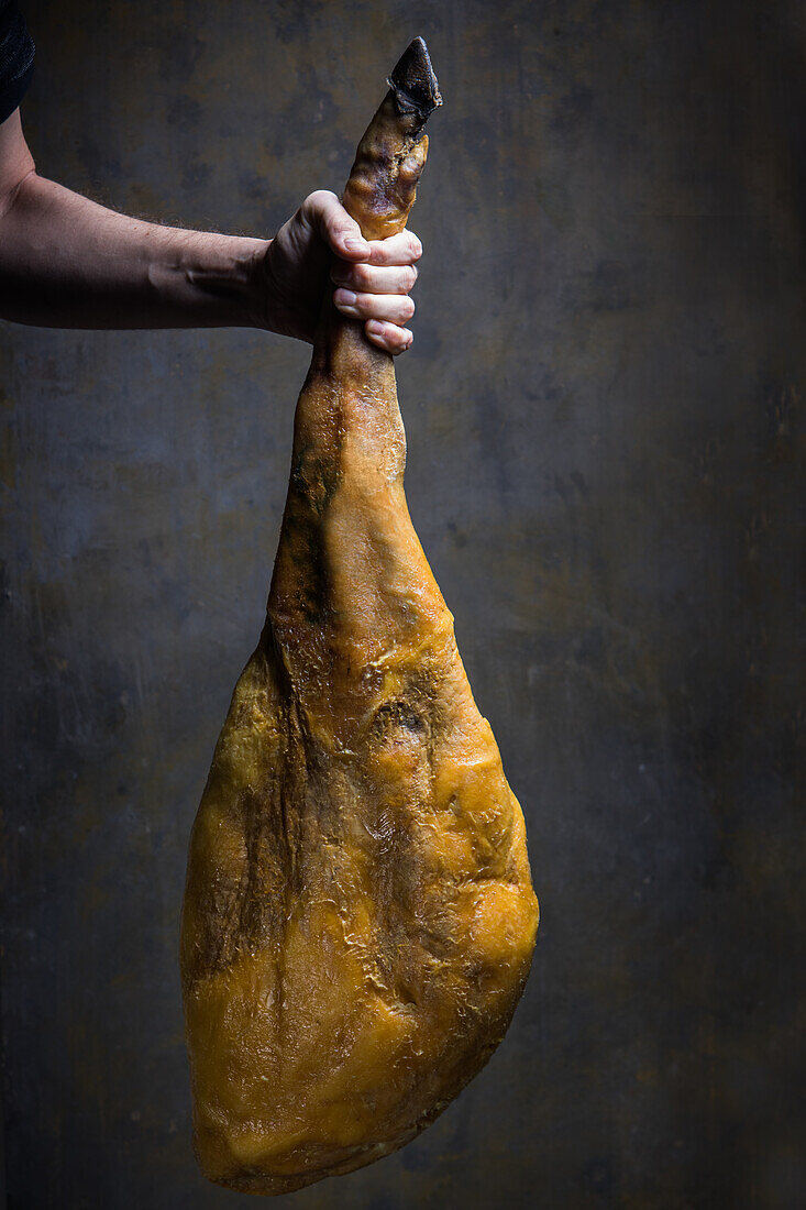 Anonymer Koch zeigt eine große trocken gepökelte iberische Schweinekeule mit goldener Haut bei Tageslicht