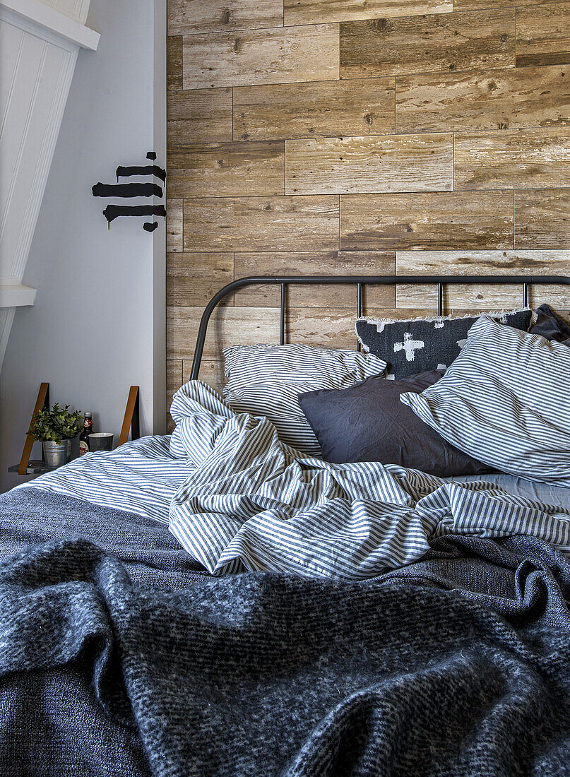 Metal bed in front of wood-panelled wall with striped bed linen, bedspread and cushions