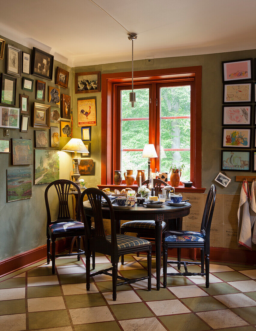 Dining area with round wooden table, picture gallery on green wall and red window frame