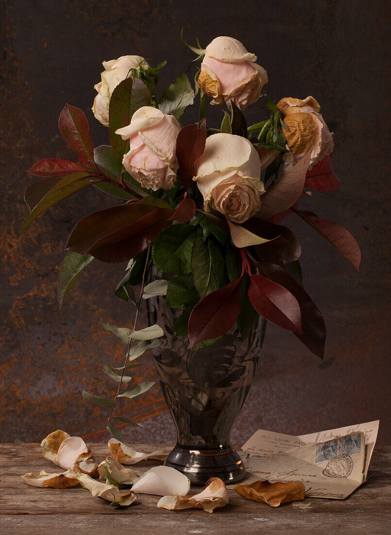 Withering pink roses and autumn leaves in a glass vase underneath an old letter